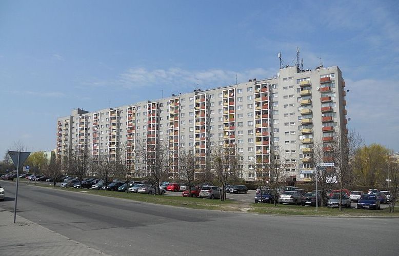 Długi wielorodzinny blok Osiedla Malinka przy ulicy Piotrkowskiej, z parkingiem i szpalerem drzew wzdłuż jezdni.