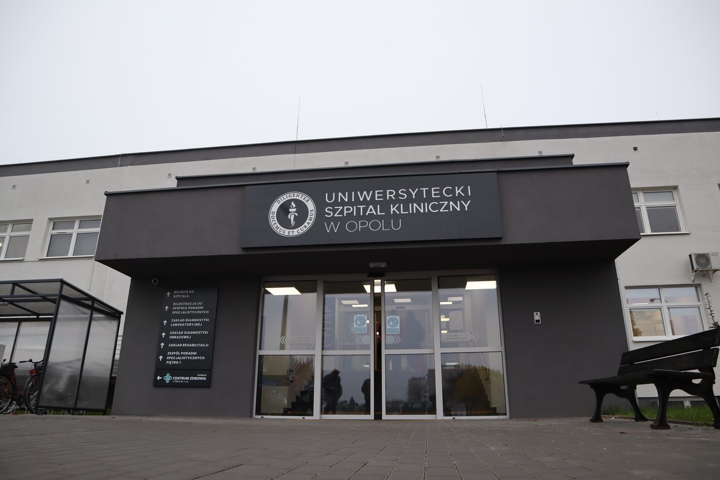 Przeszklone wejście główne Uniwersyteckiego Szpitala Klinicznego w Opolu z ciemną elewacją i nazwą szpitala nad drzwiami.