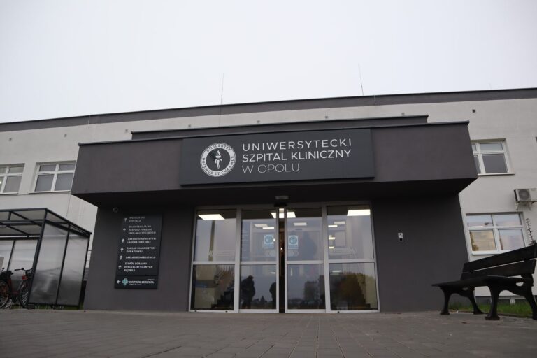 Przeszklone wejście główne Uniwersyteckiego Szpitala Klinicznego w Opolu z ciemną elewacją i nazwą szpitala nad drzwiami.